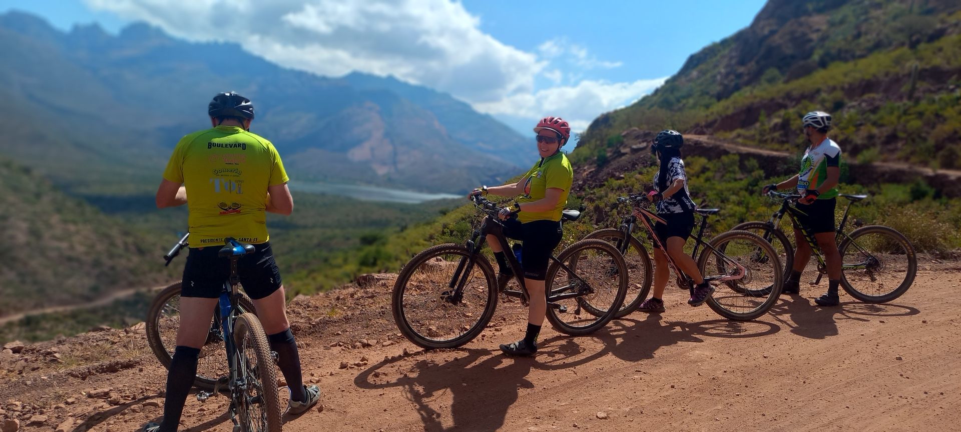 Cuatro ciclistas en bicicletas de montaña se detienen en un camino de tierra, mirando una cadena montañosa y la laguna de Brealito