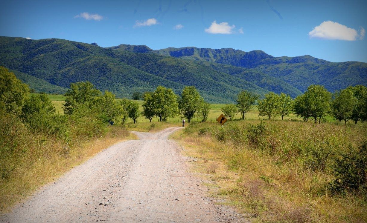 Camino de tierra que conduce a montañas, árboles verdes y un cielo azul.