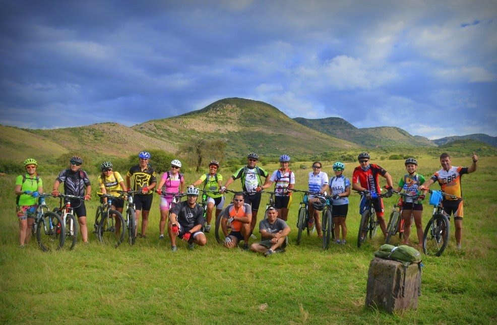 Grupo de ciclistas posando en un campo con un fondo montañoso; cielo nublado.