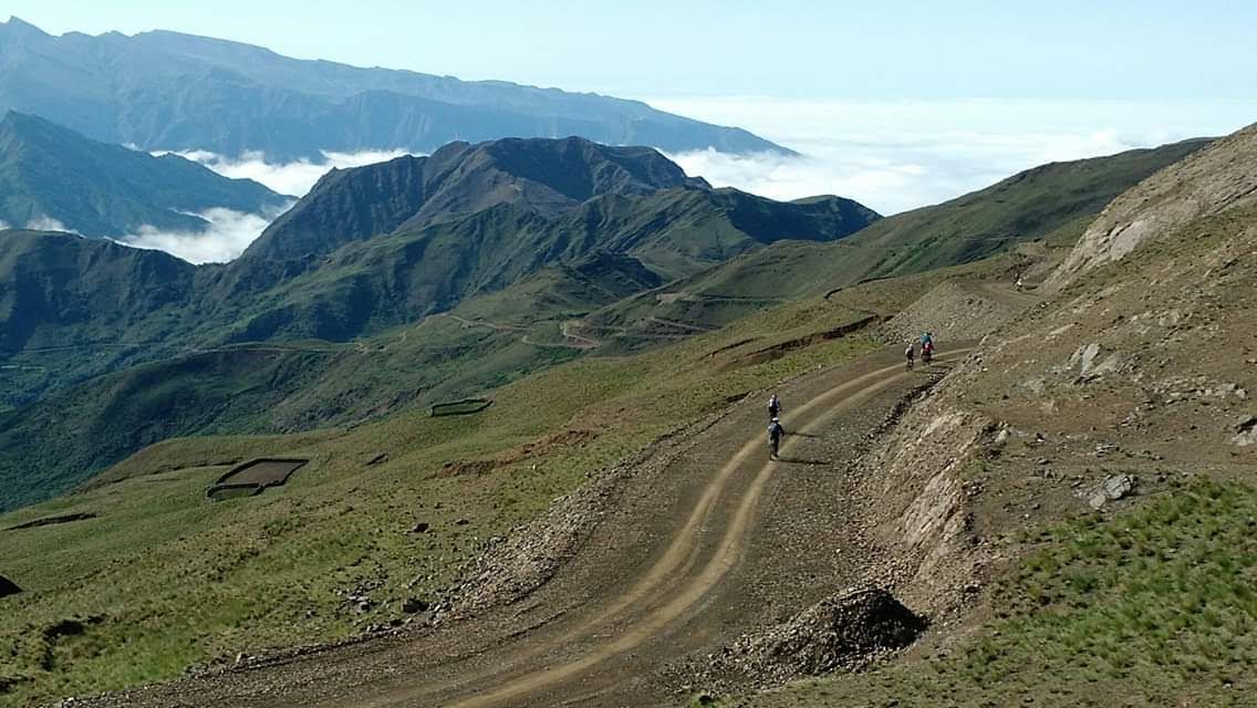 Carretera de montaña con ciclistas, colinas verdes y valle nublado.
