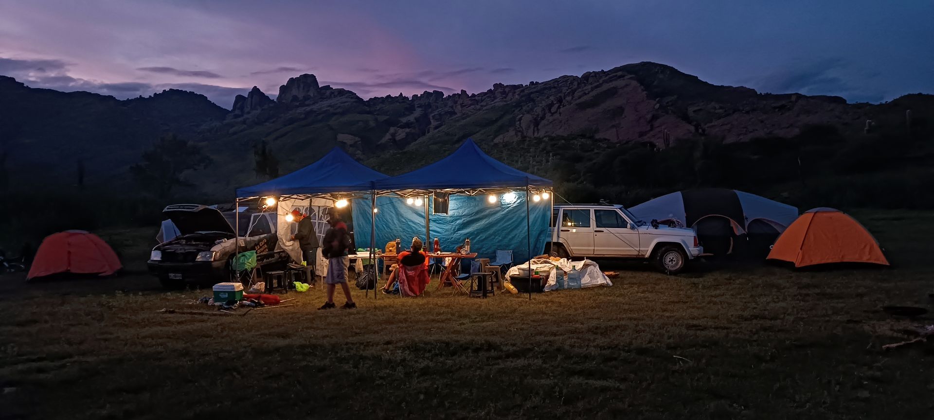 Acampada al anochecer con tiendas de campaña, coches y un grupo reunido alrededor de luces. Montañas al fondo.