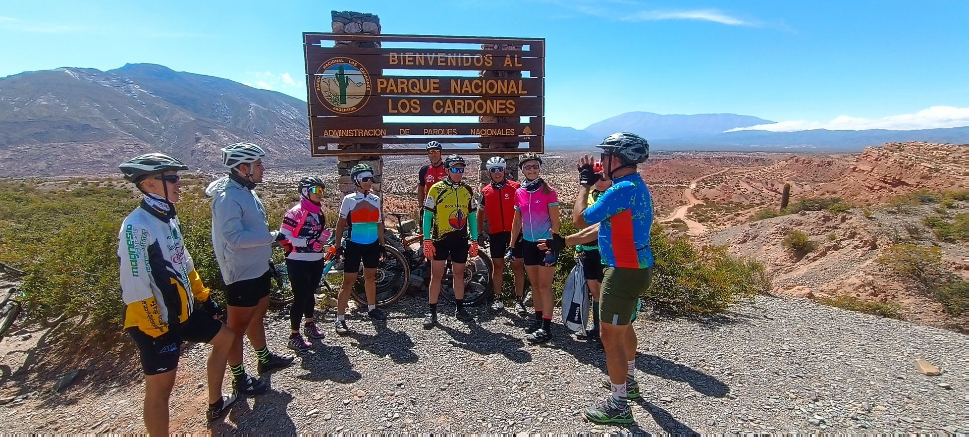 Ciclistas posan junto a un cartel que indica el Parque Nacional Los Cardones, en un paisaje montañoso.