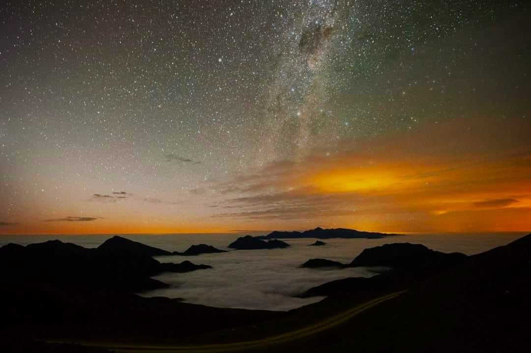 Vía Láctea sobre montañas recortadas, niebla y un horizonte iluminado de color naranja.