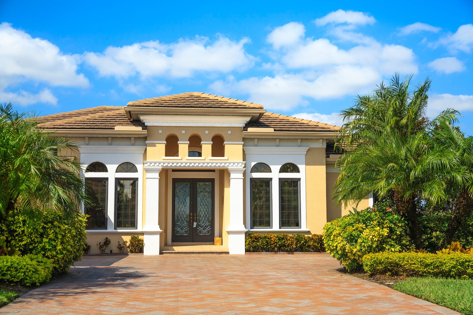 Tan stucco house with brick driveway, arched windows, and palm trees under a blue sky.