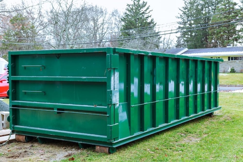 A large green dumpster is sitting in the grass in front of a house.