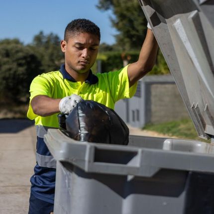 A man in a yellow shirt is putting a black bag into a trash can