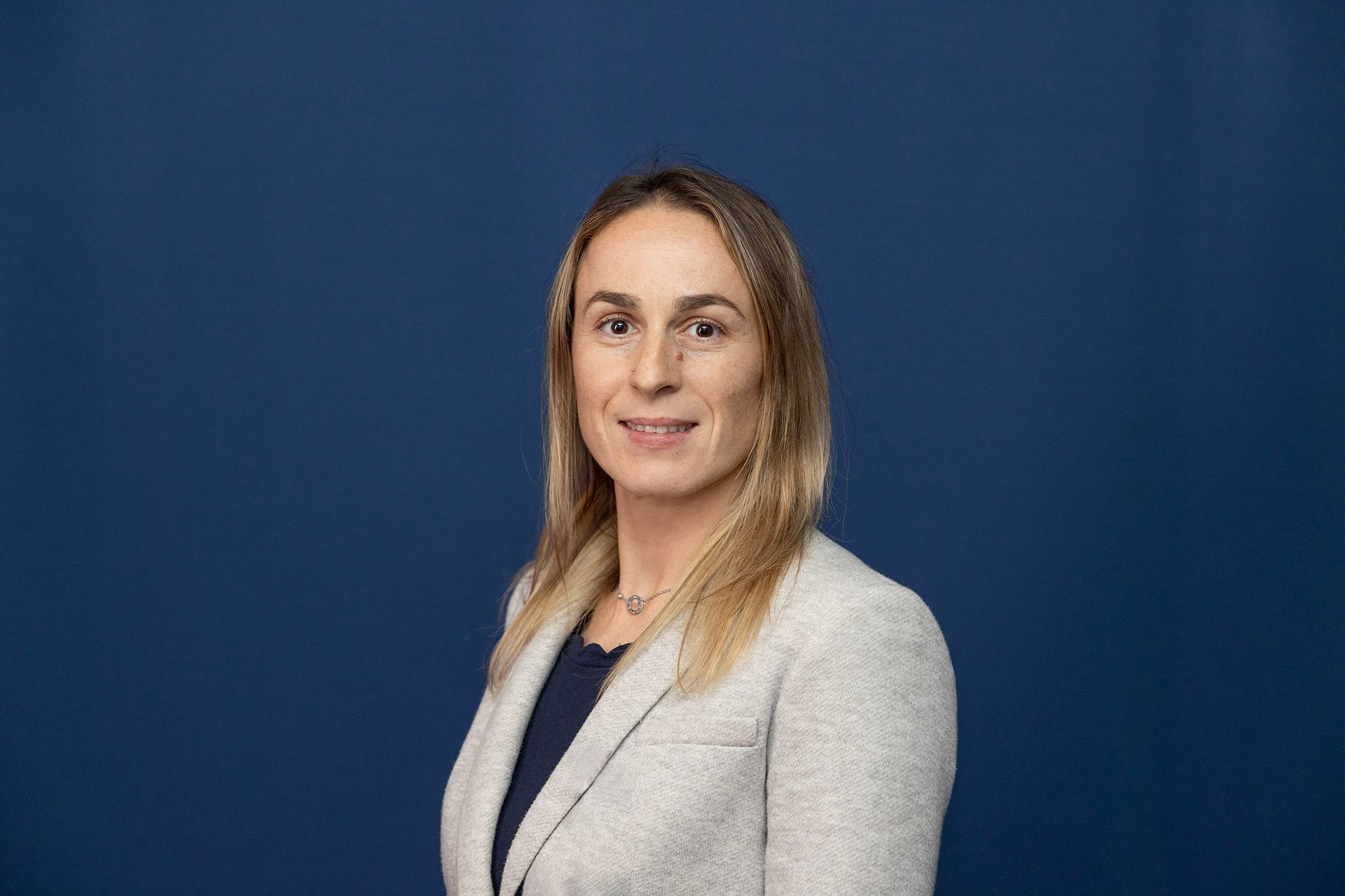 Woman with blonde hair wearing a blazer, smiling against a blue background.