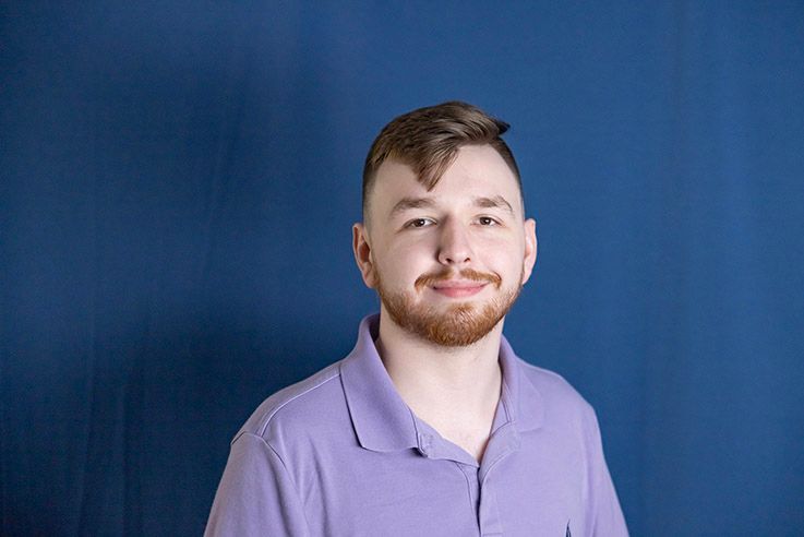 Man with short brown hair and beard, wearing a purple polo shirt, smiles in front of a blue background.