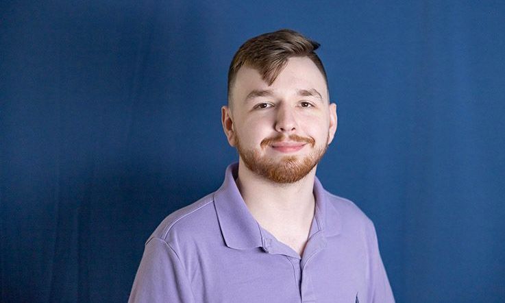 Man with brown hair and beard smiles in purple shirt against blue background.