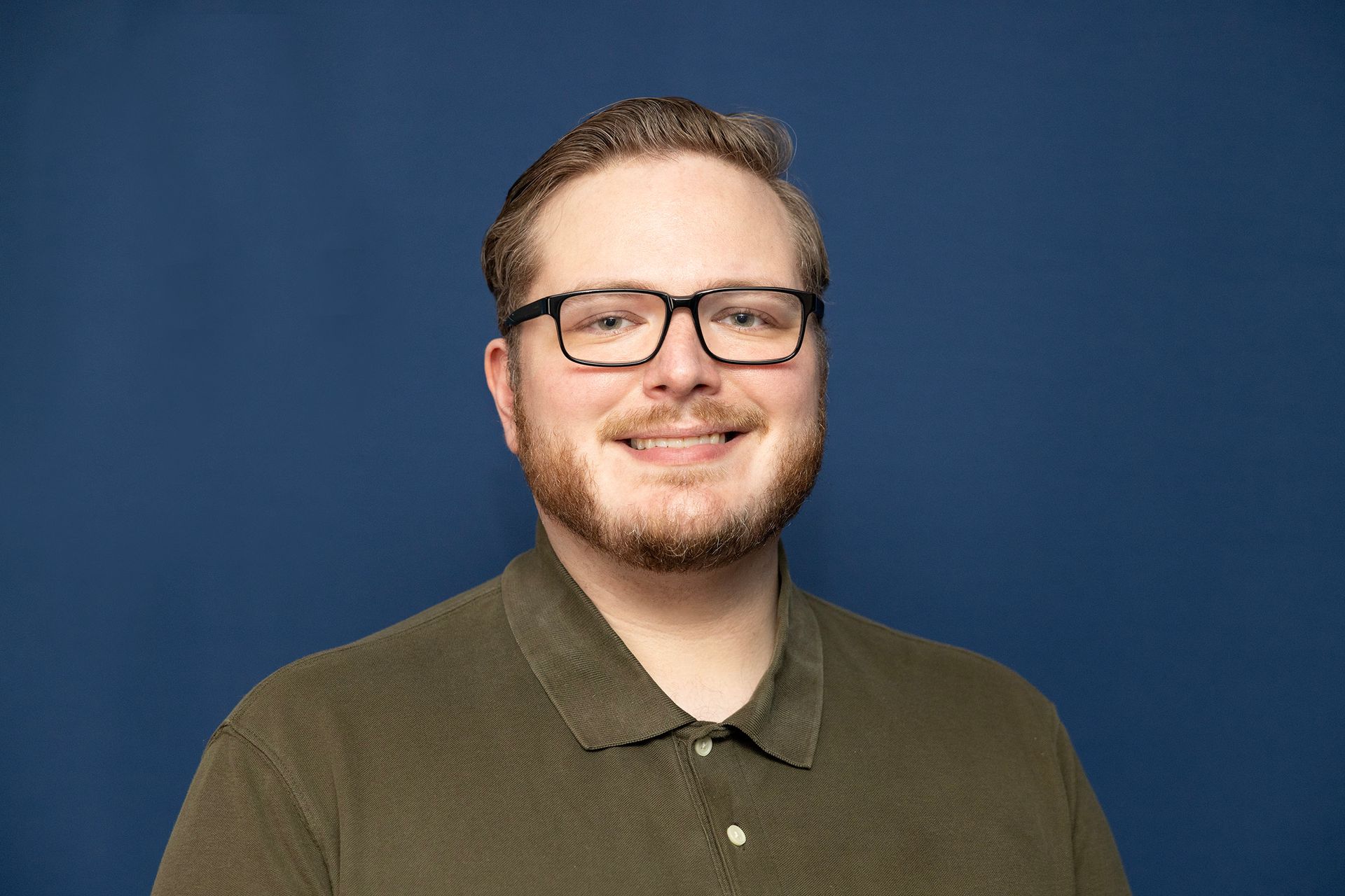 Man with beard, glasses, smiling, in green shirt, against blue background.