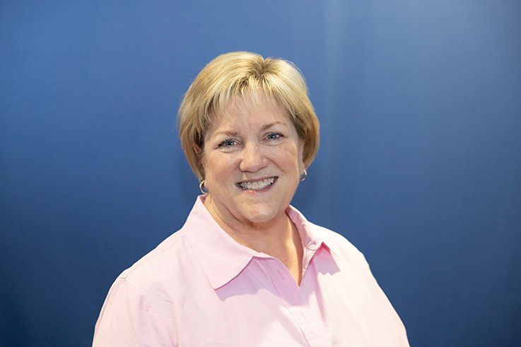 Woman with blonde hair, smiling, wearing a pink button-down shirt, in front of a blue backdrop.