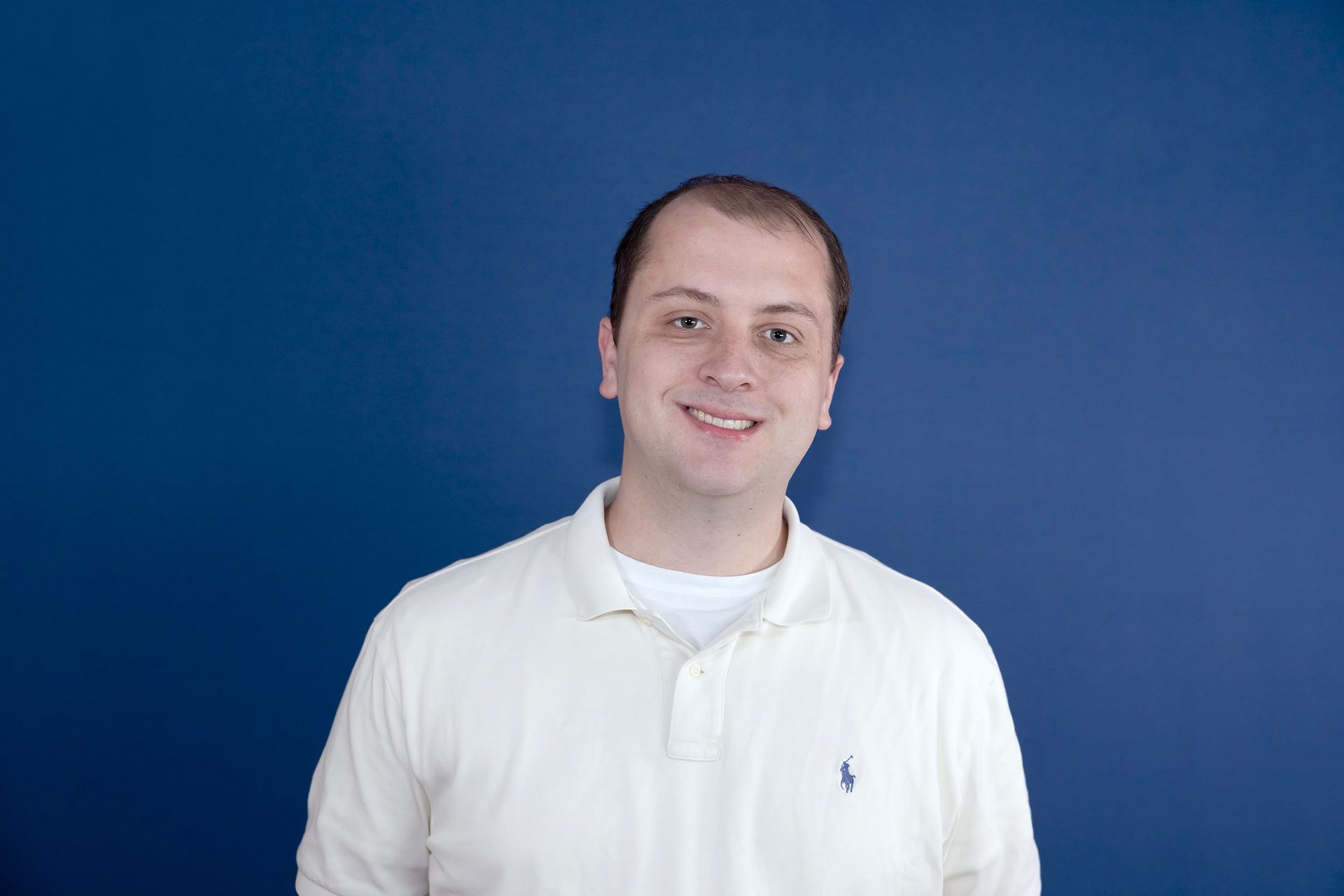 A man in a white polo shirt is smiling in front of a blue wall.