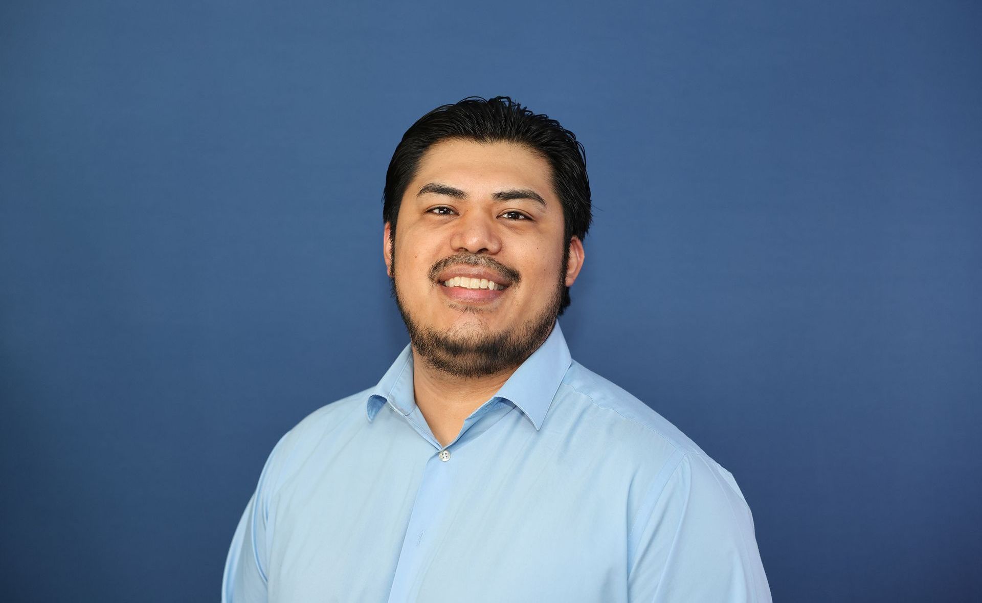 Man with dark hair and beard smiles broadly in light blue shirt against blue backdrop.