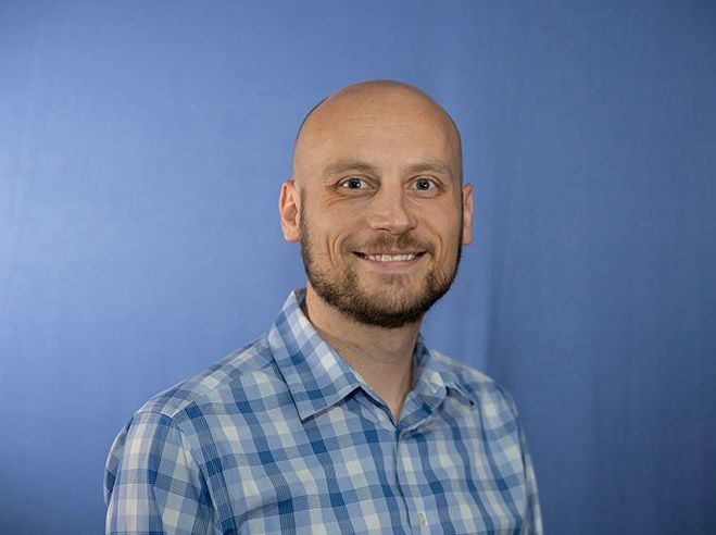 Bald man with beard, smiling, in blue checkered shirt, against blue background.
