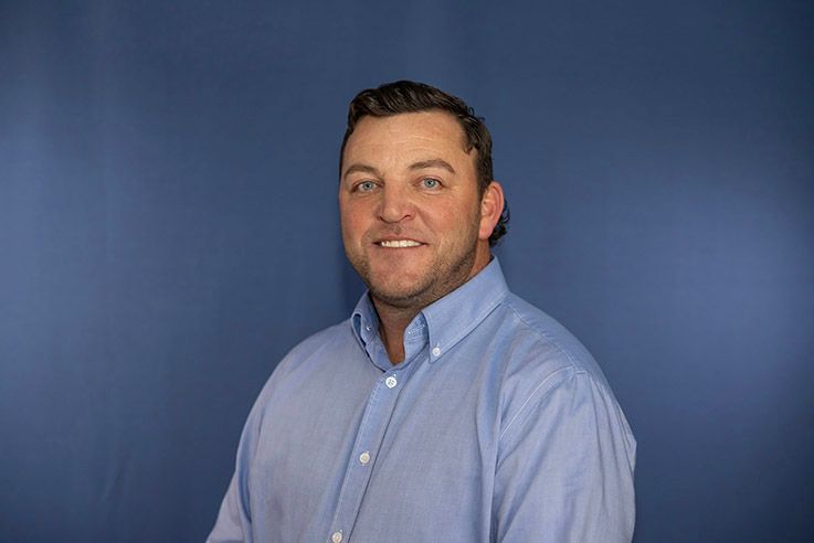 Man in light blue shirt smiles in front of a solid blue backdrop.