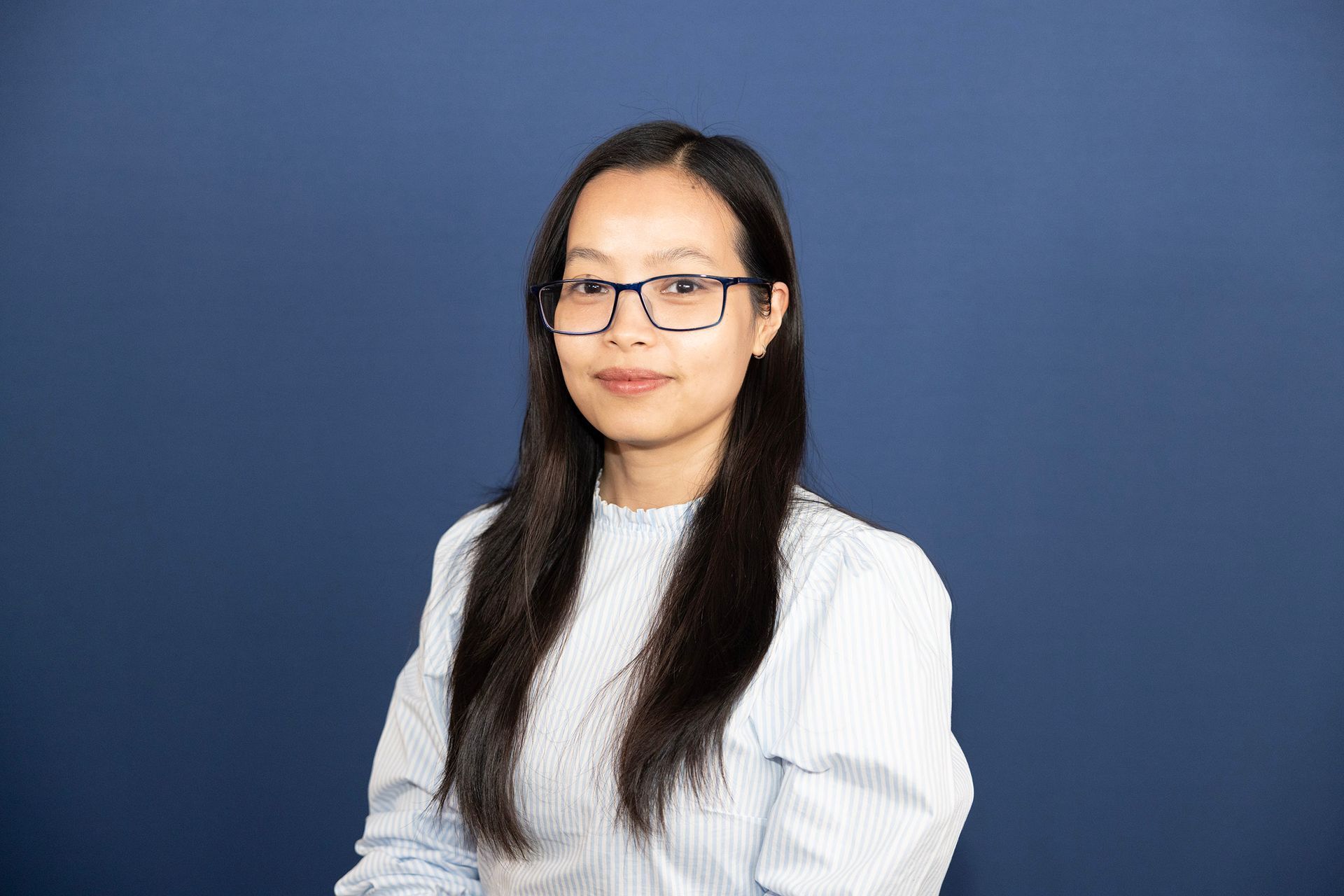 A woman wearing glasses and a white shirt is standing in front of a blue background.