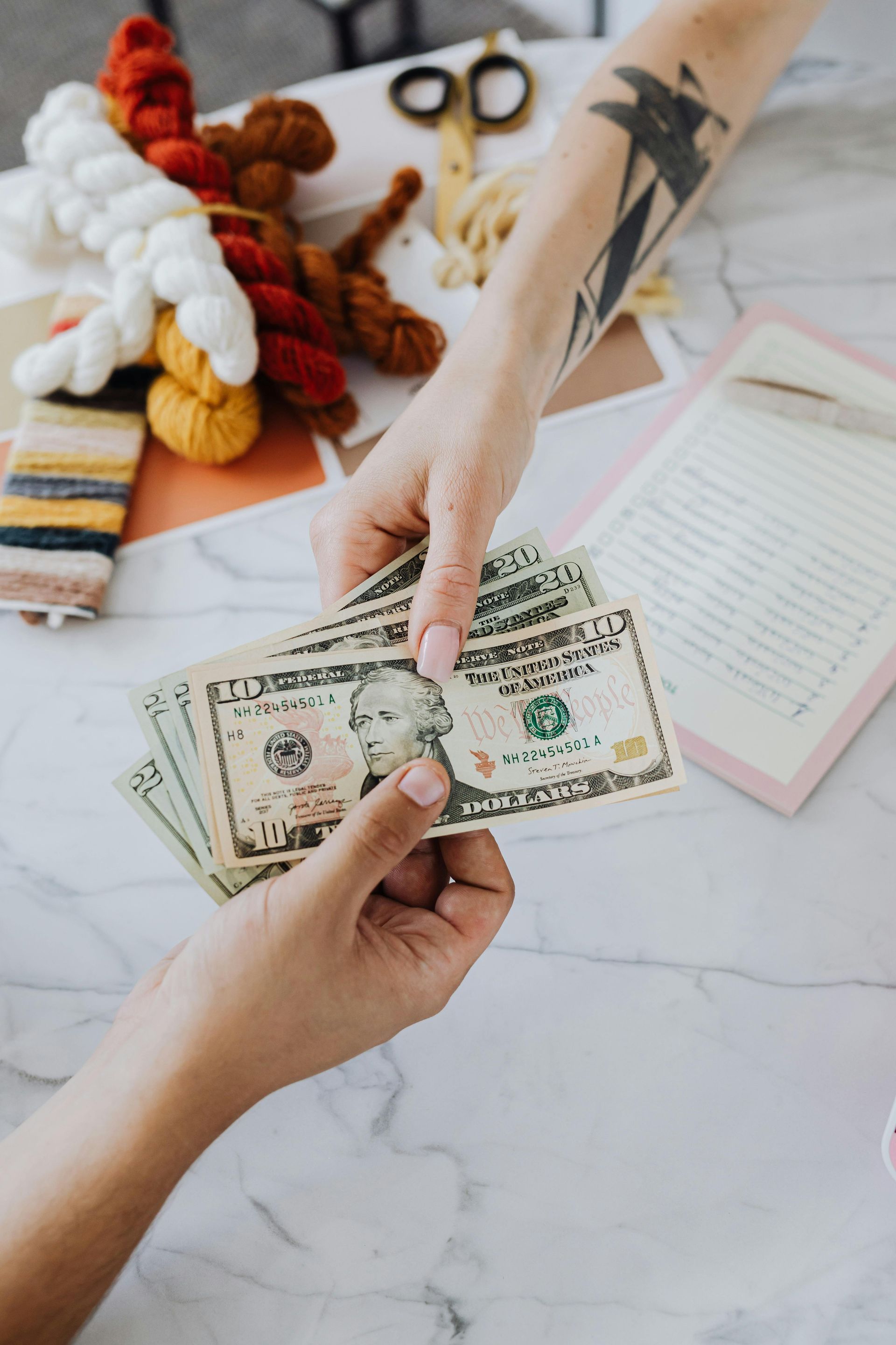 Hands exchanging US dollar bills over a white table with crafting supplies.