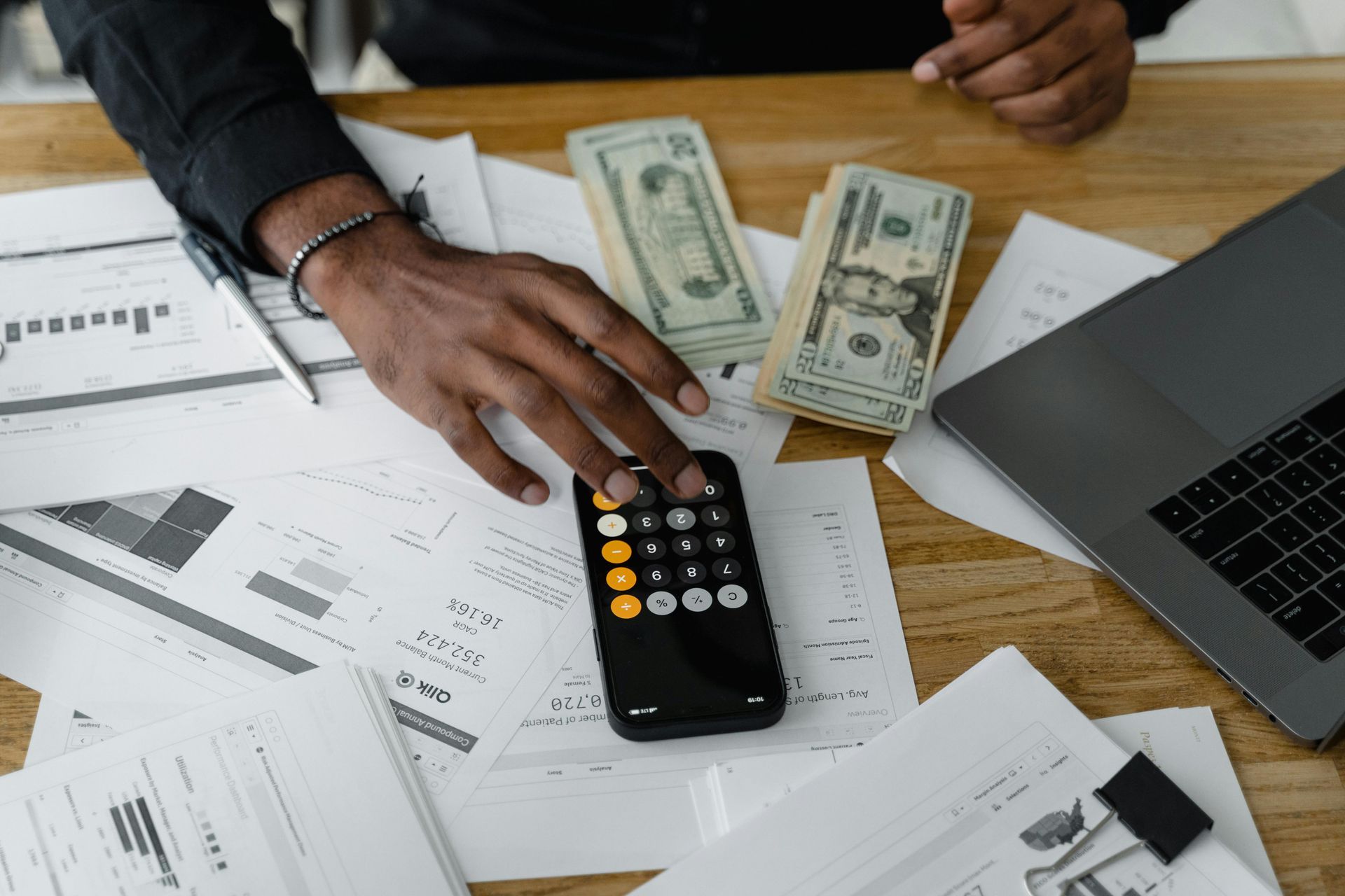 Person using a calculator, with cash, laptop, and papers on a desk, possibly financial analysis.