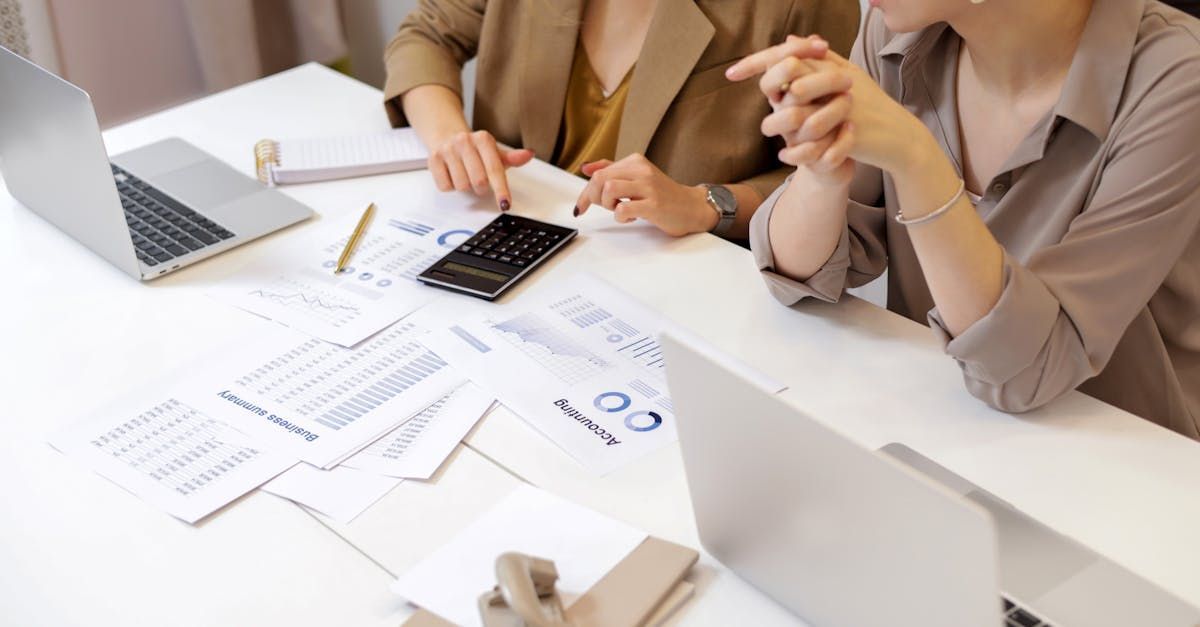 Two women are sitting at a table with laptops and papers.