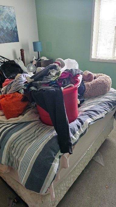 A bedroom with a bed filled with clothes and a red bin on top of it.