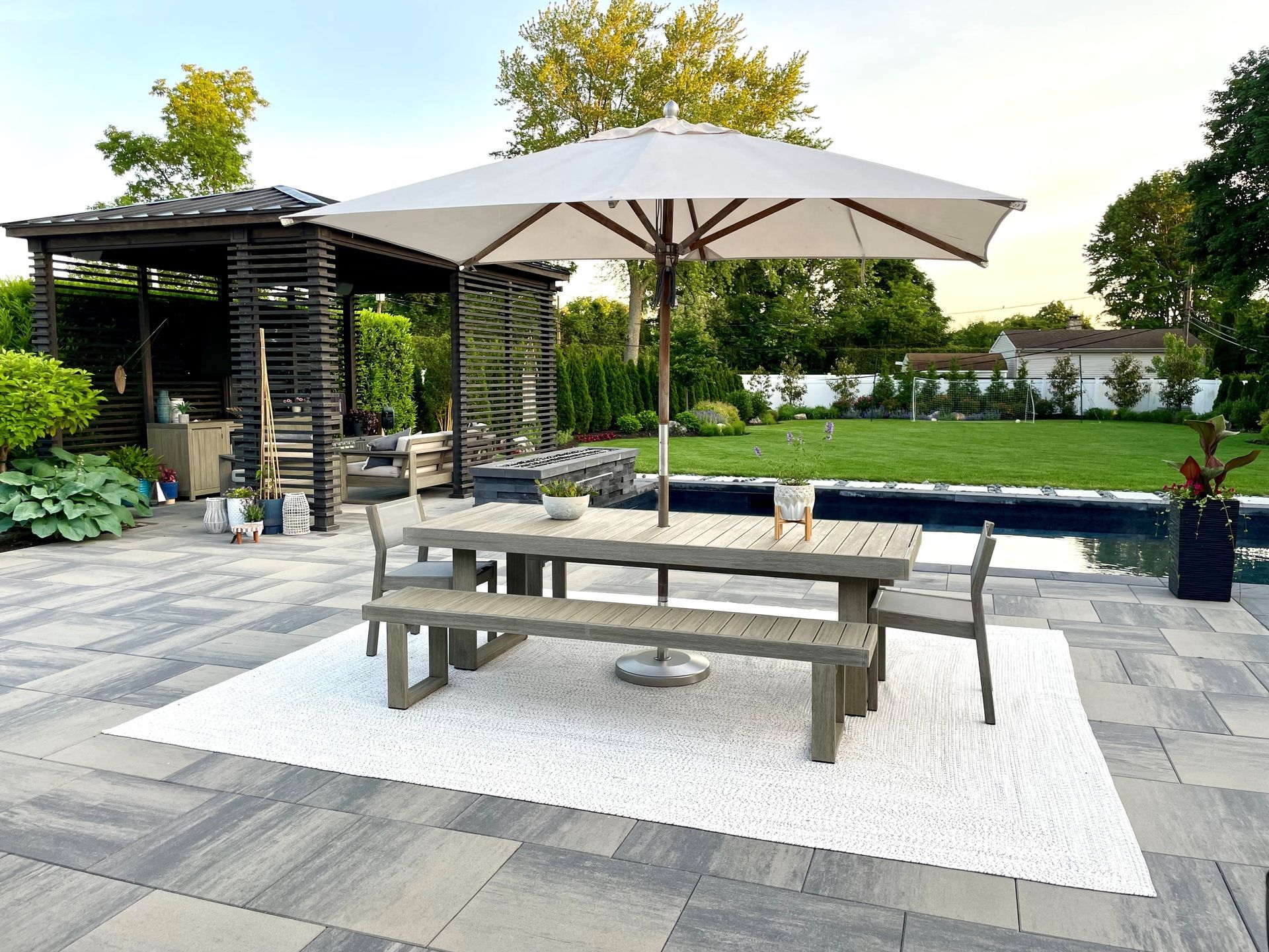 Backyard patio with picnic table, umbrella, chairs, and pergola beside a lawn and garden