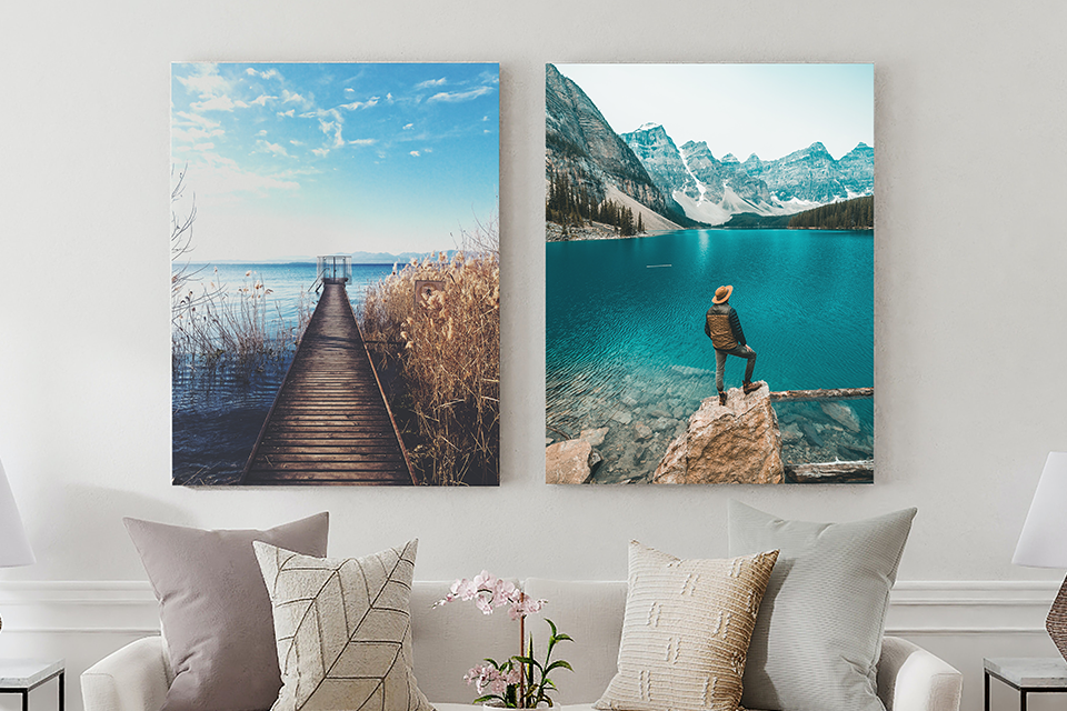 Two metal prints, a dock on a waterscape, & a man in a hat in front of a lake & mountains, hang on a living room wall.