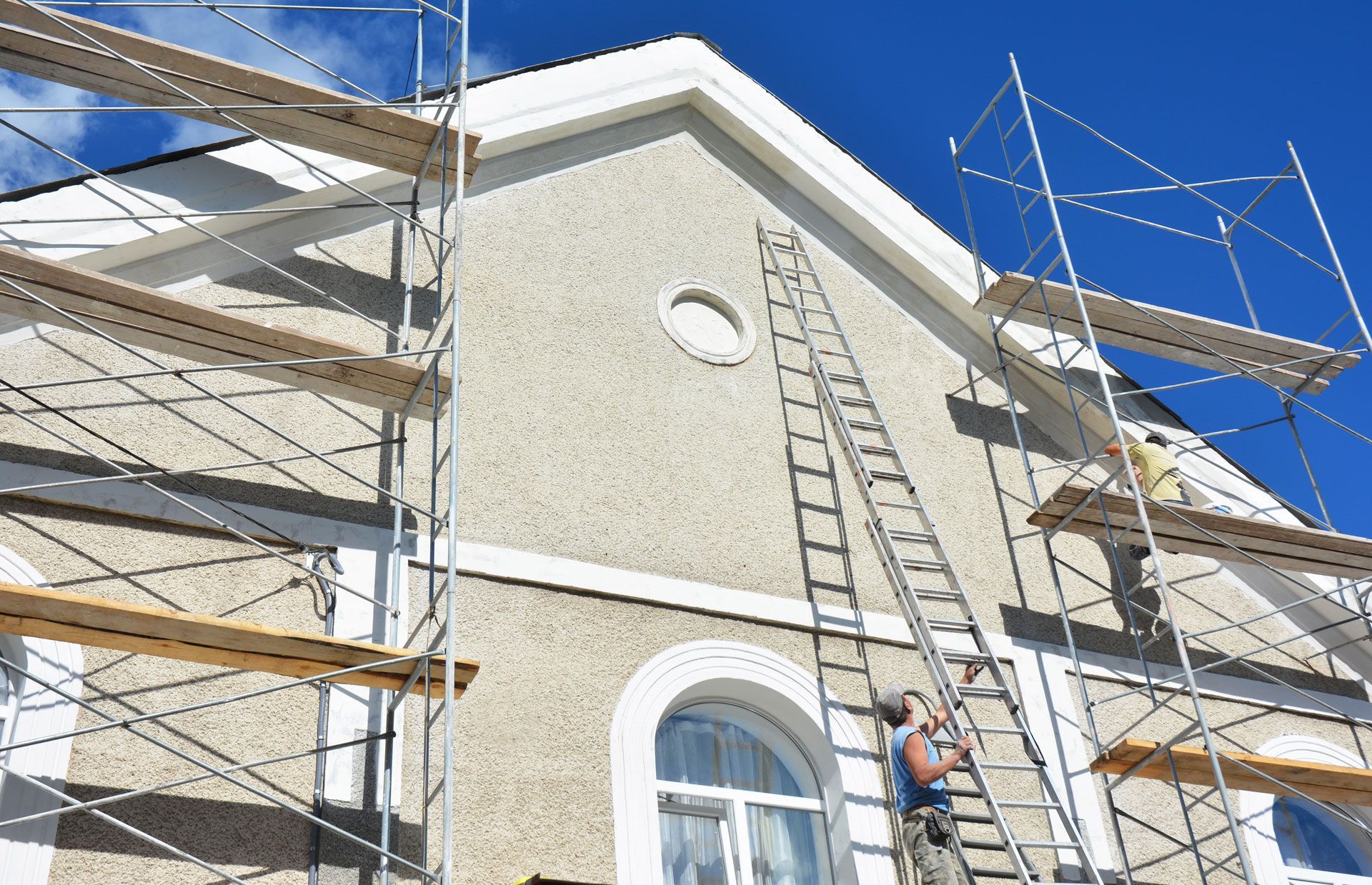 Building exterior under construction; scaffolding, ladder, worker. Blue sky.