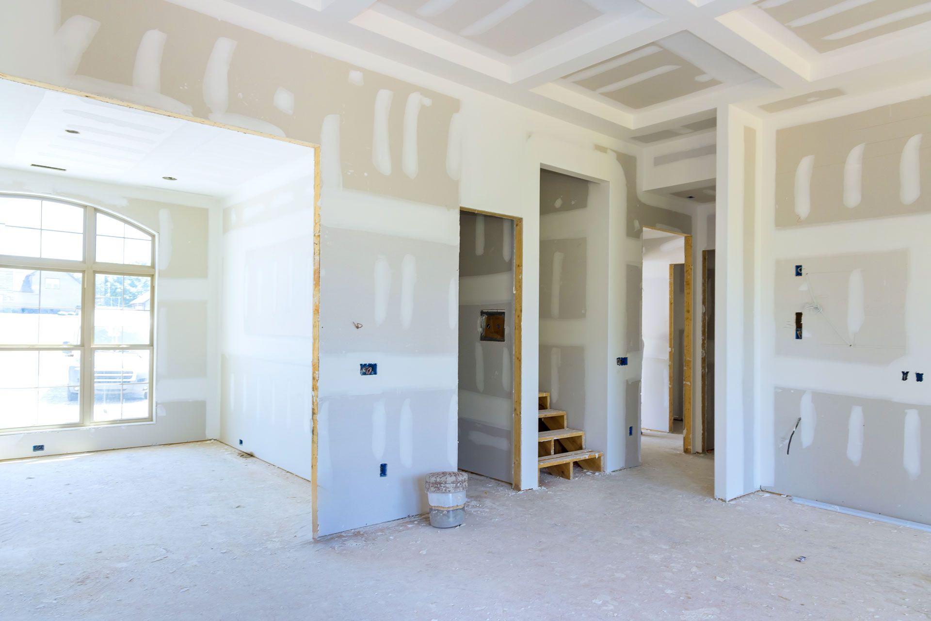 Interior of a room under construction, with drywall walls and ceiling, bare stairs, and an arch window.
