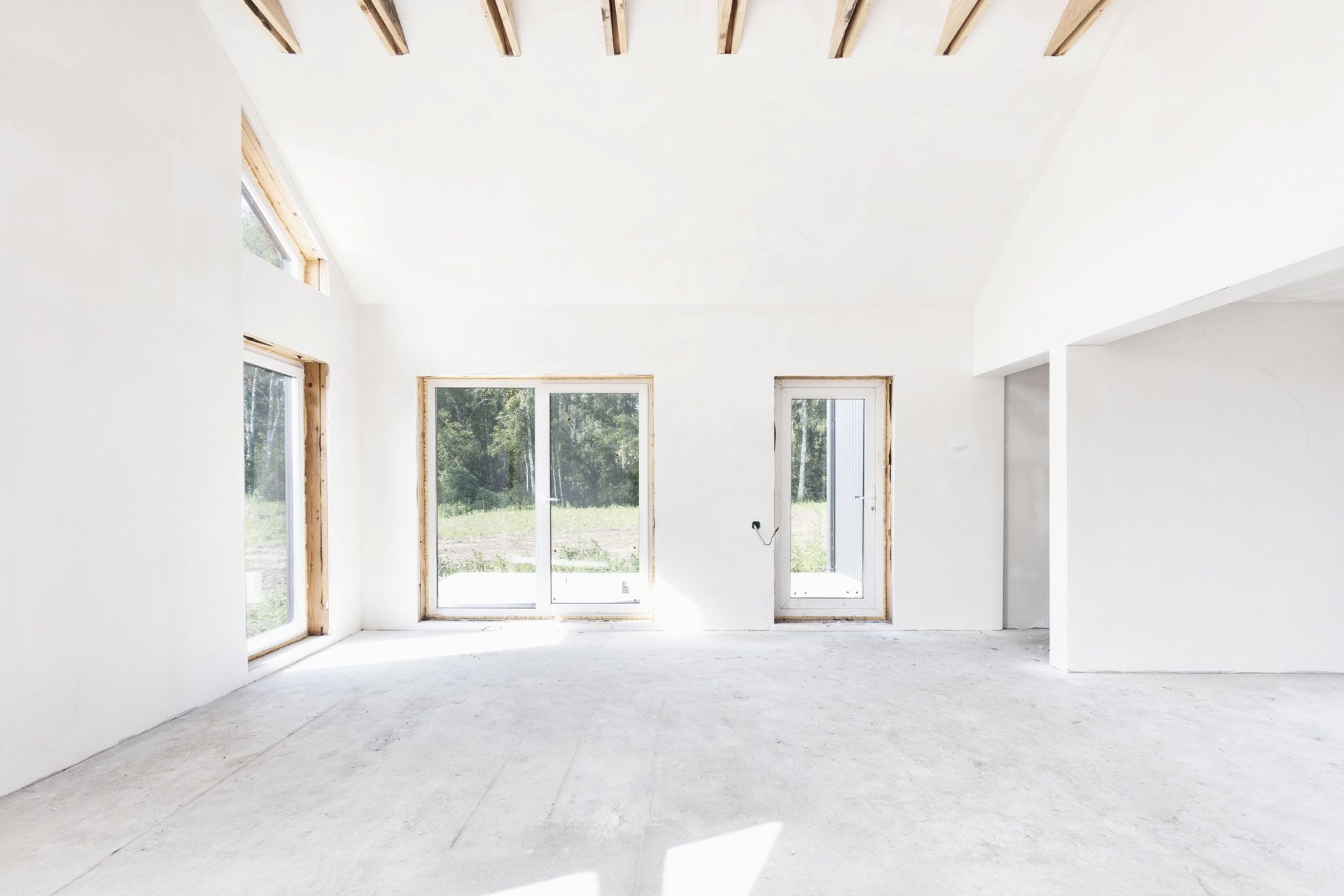 Empty, bright white room with large windows and a concrete floor. Wooden beams on the ceiling.