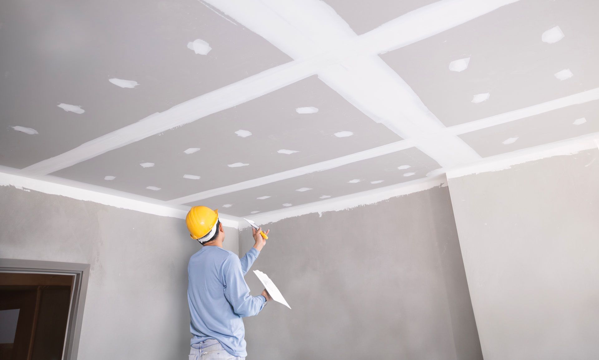 Person in a hard hat applying joint compound to a ceiling with a trowel, during construction.