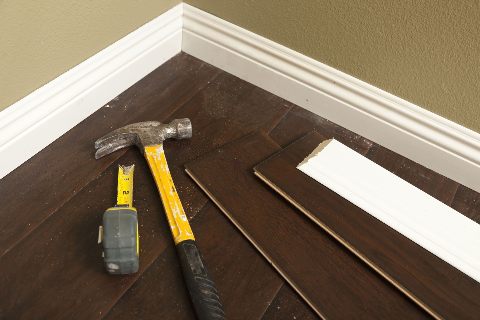 Hammer, measuring tape, and flooring materials on dark wood floor, next to white molding.