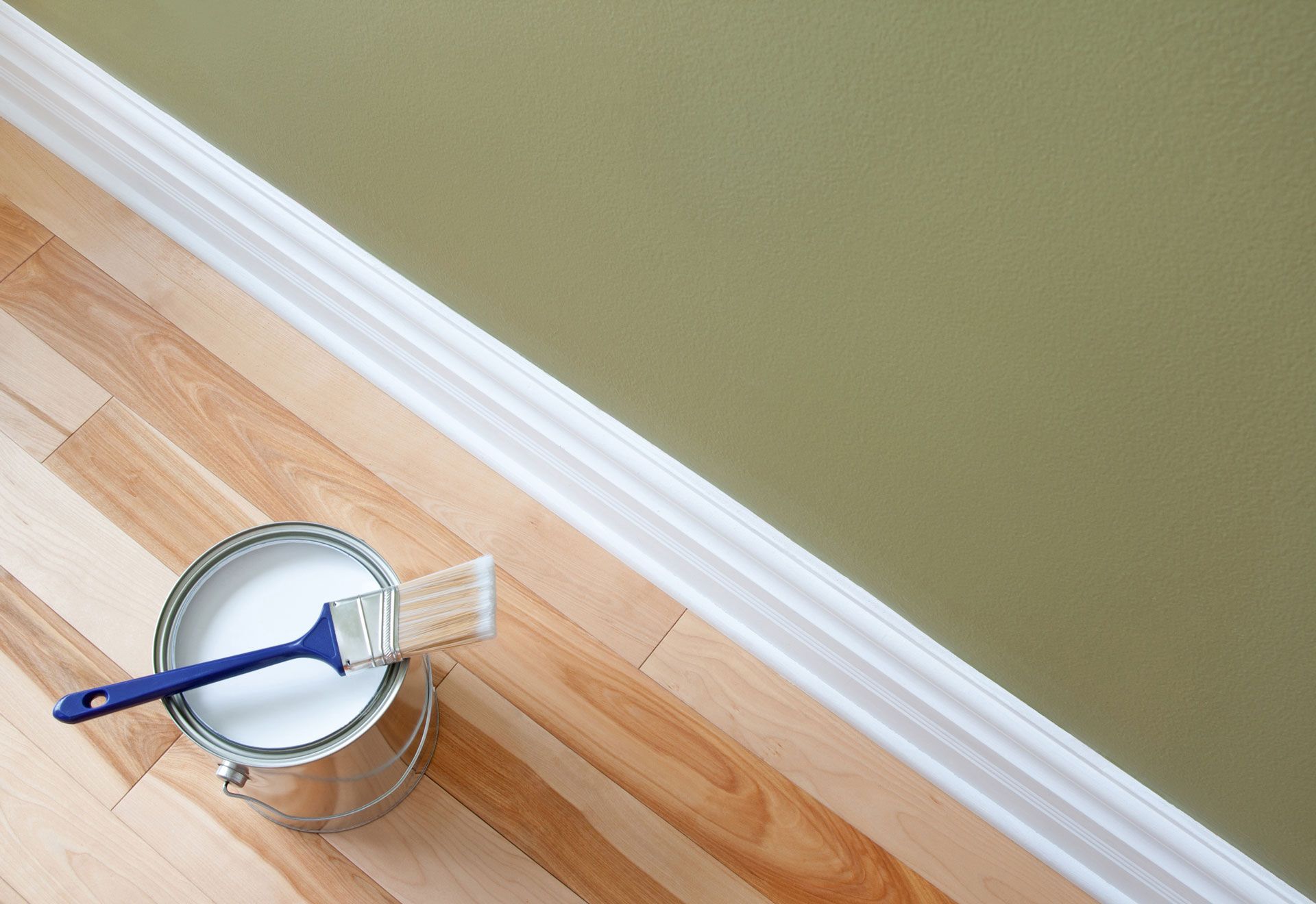 Paint can with paintbrush on hardwood floor beside green painted wall.