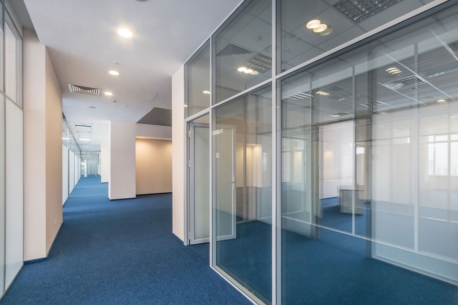 Empty office hallway with blue carpet, glass-walled rooms, and white walls and ceilings.