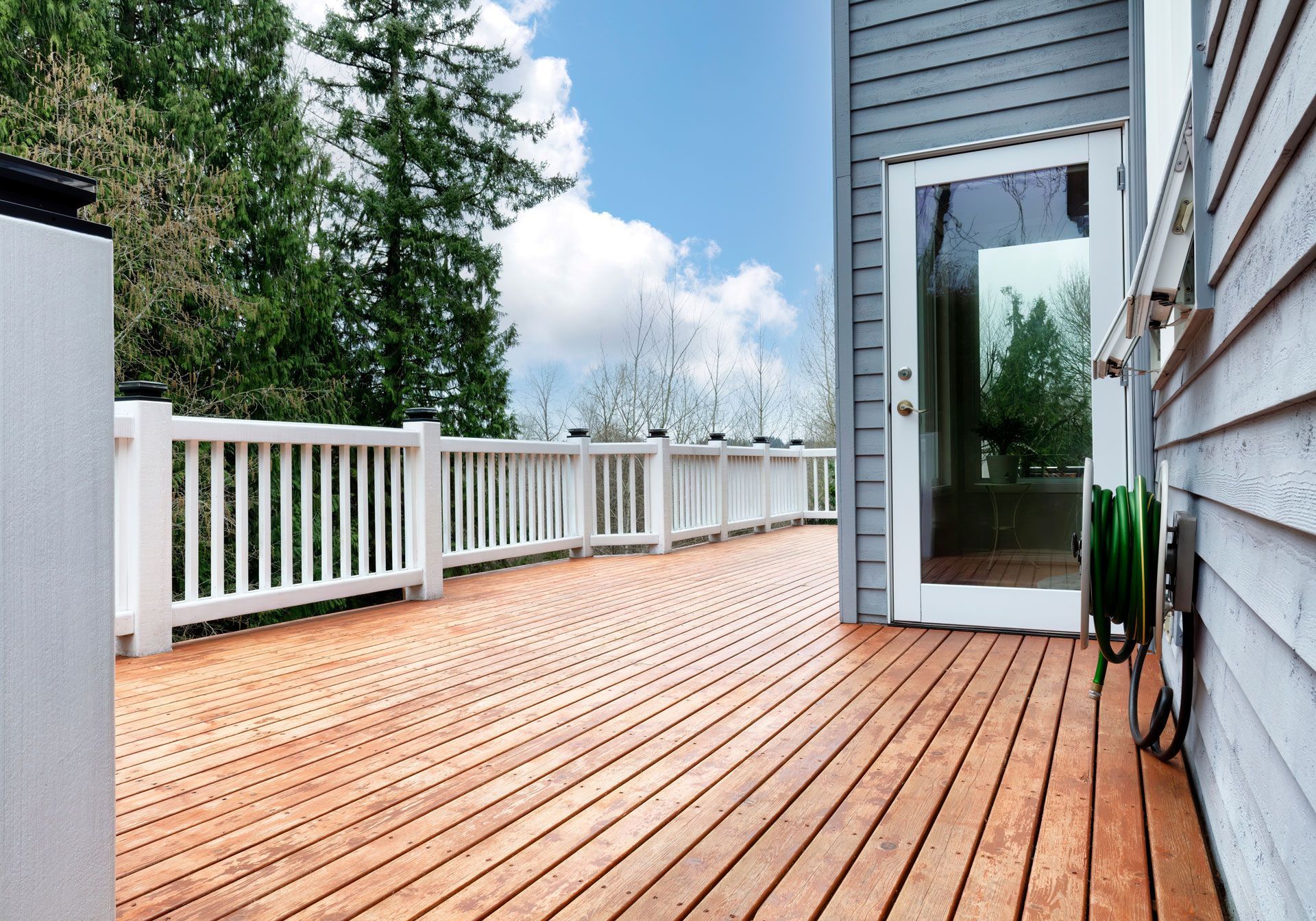 Wooden deck with white railing, glass door, and green hose on the side of a gray house, with trees in the background.