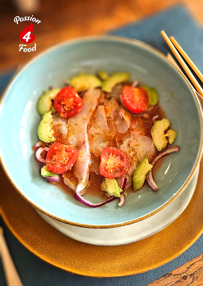 In een blauwe kom ligt een verse visceviche met plakjes cherrytomaatjes, avocado en rode ui, geserveerd met eetstokjes.