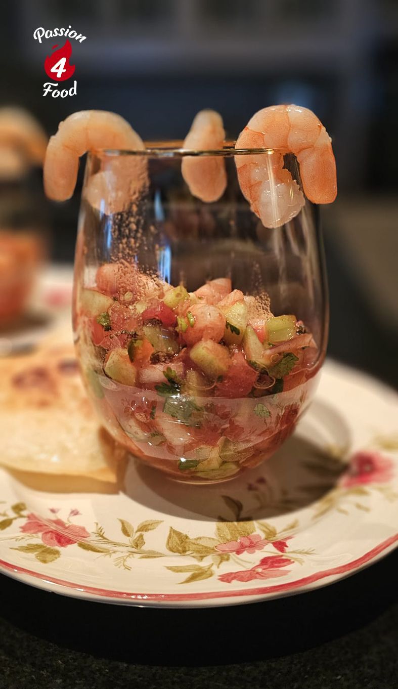Een glas gevuld met een kleurrijke garnalensalsa als voorgerecht, gegarneerd met drie garnalen, geserveerd op een bord met bloemmotief.
