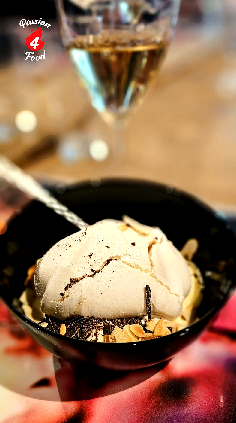 Ile Flottante in een zwarte kom, besprenkeld met chocoladesaus, met een glas op de achtergrond.