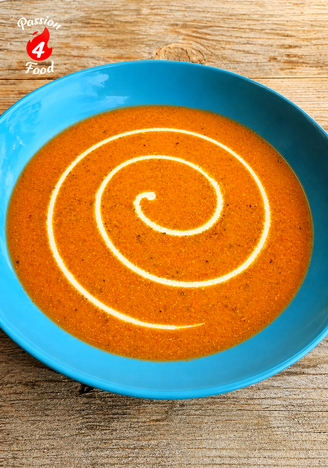 Paprika-Courgette soep in een blauwe kom met een witte roomkrul op een houten tafel.