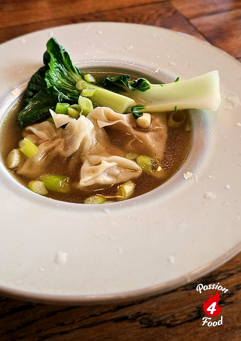 Een witte kom wontonsoep met bladgroenten en mals vlees op een houten tafel.