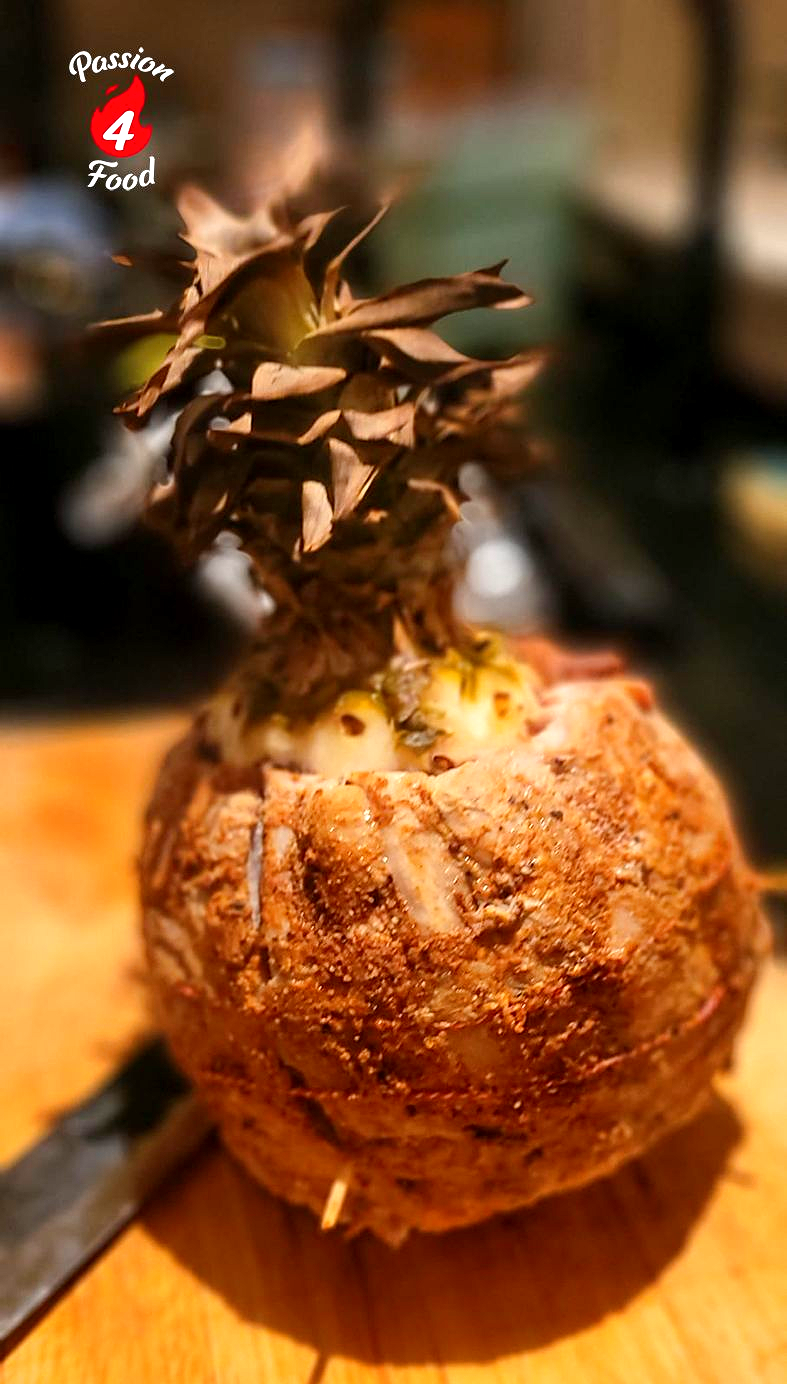 Een kleine ananas, met het loof er nog aan, ligt op een houten plank, met een wazige keukenachtergrond.