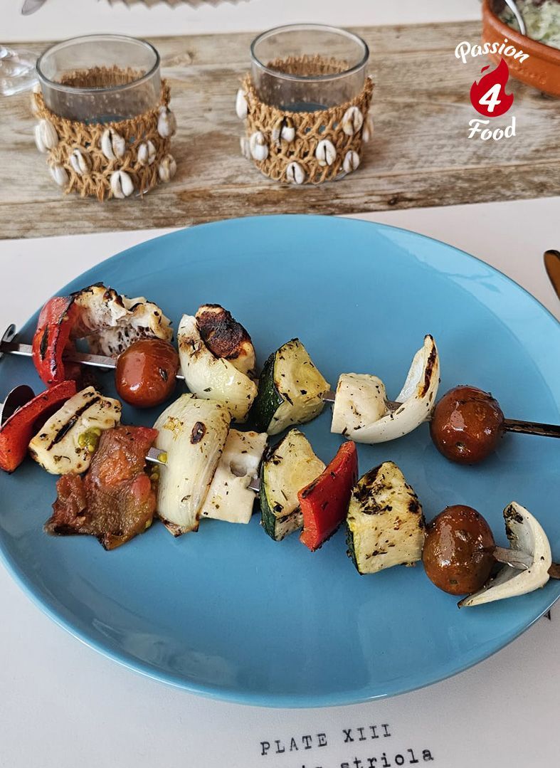 Een blauw bord met gegrilde haloumi en groenten op een tafel, met twee glazen op de achtergrond.