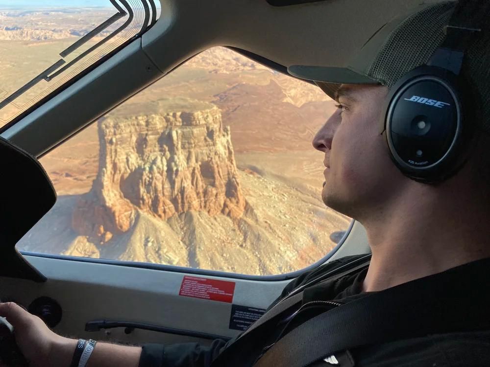 Pilot in headset flying over a desert canyon at sunset, seen through the cockpit window.