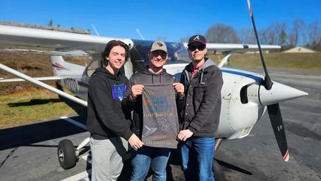Three people posing beside a small airplane, holding a decorative sign on a sunny tarmac.