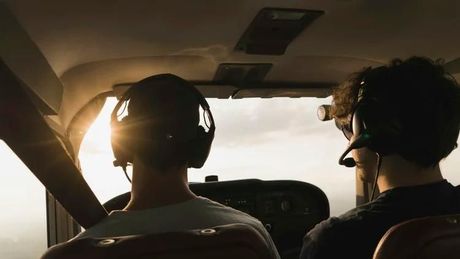 Two headset-wearing people sit in a small aircraft, looking toward the bright cockpit window.