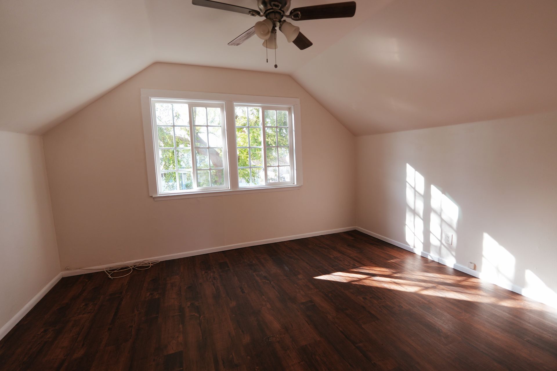 Empty attic room with dark wood floors, sloped ceiling, and sunlight through a window.