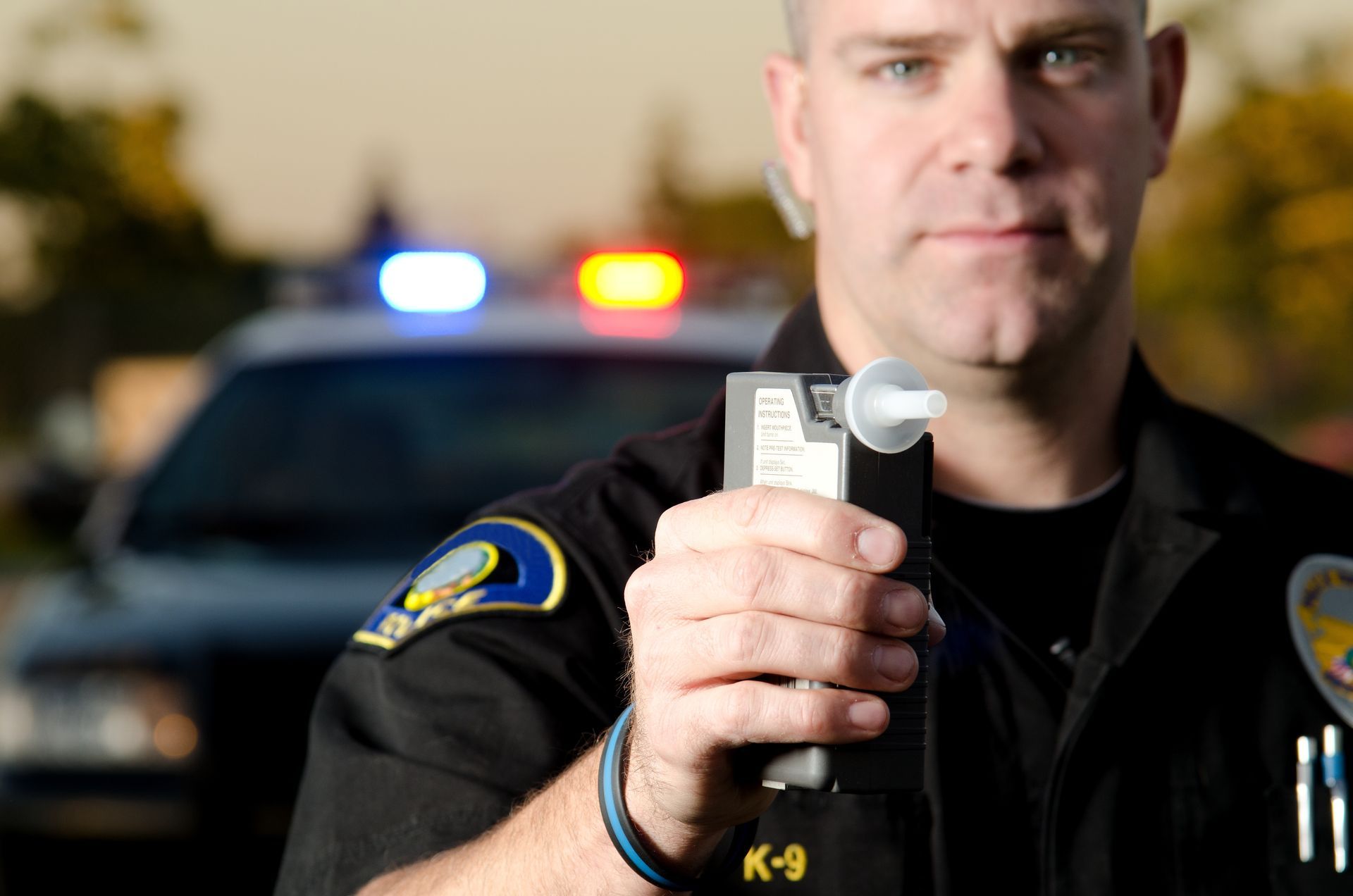 A police officer conducting a roadside breathalyzer test with patrol car lights in the background. A police officer conducting a roadside breathalyzer test with patrol car lights in the background.