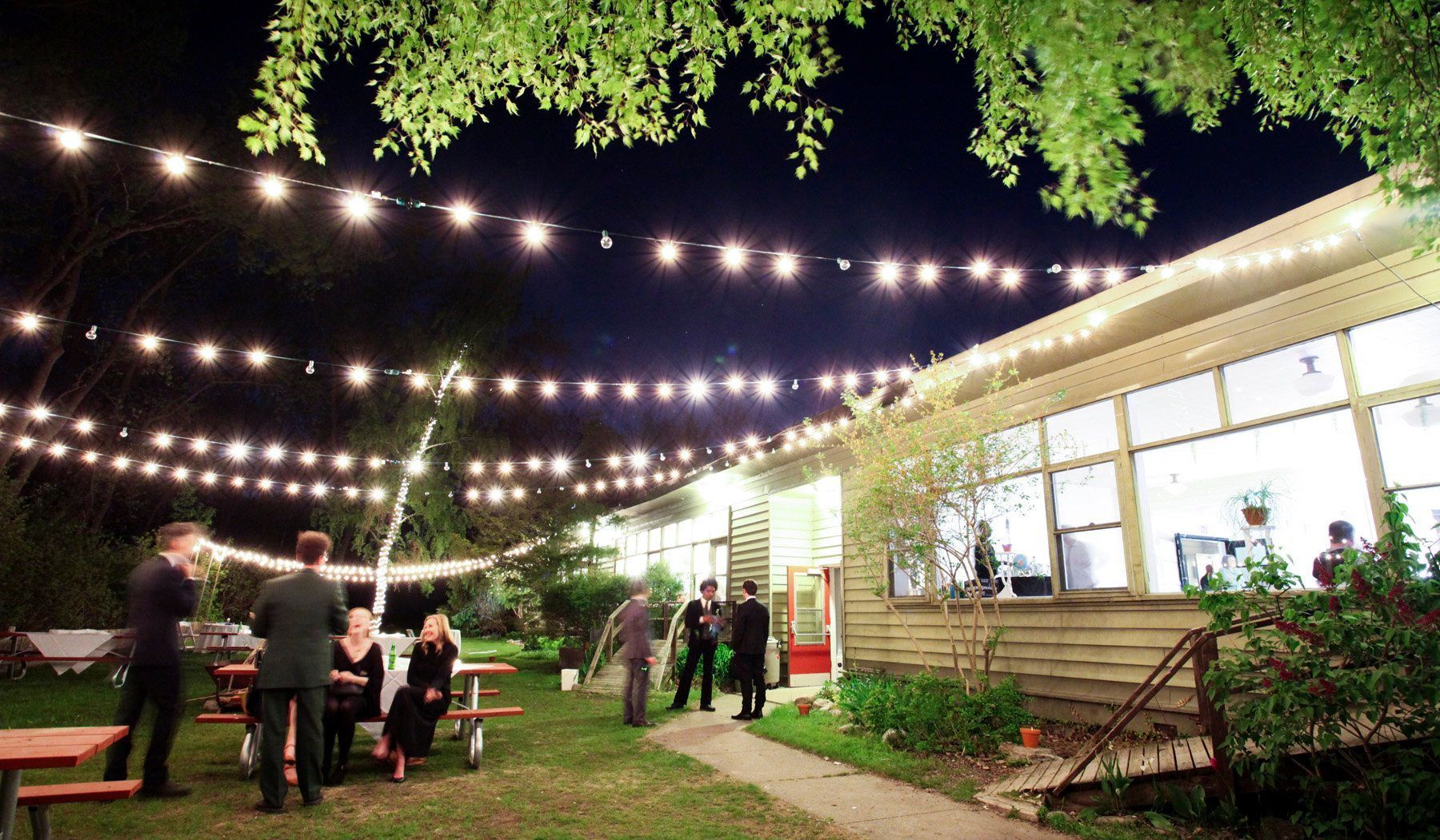 Outdoor evening gathering with string lights, guests near a wooden building and picnic tables.