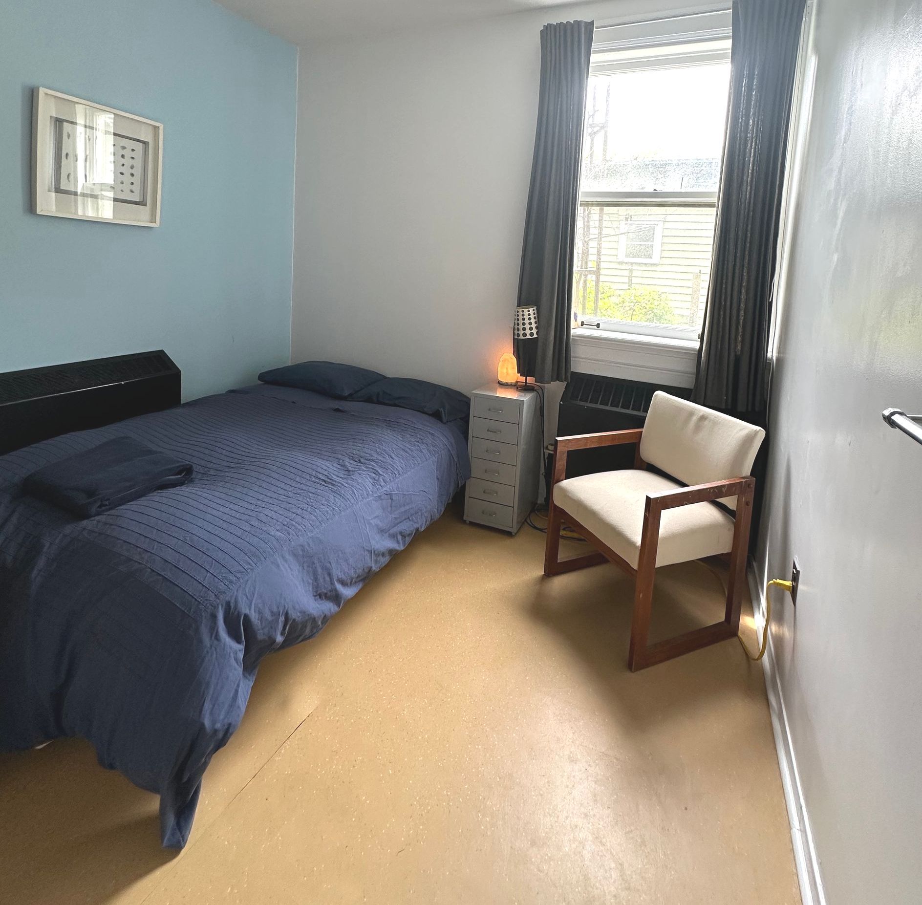 Small bedroom with bed, chair, and window, featuring blue walls, dark bedding, and gray curtains.