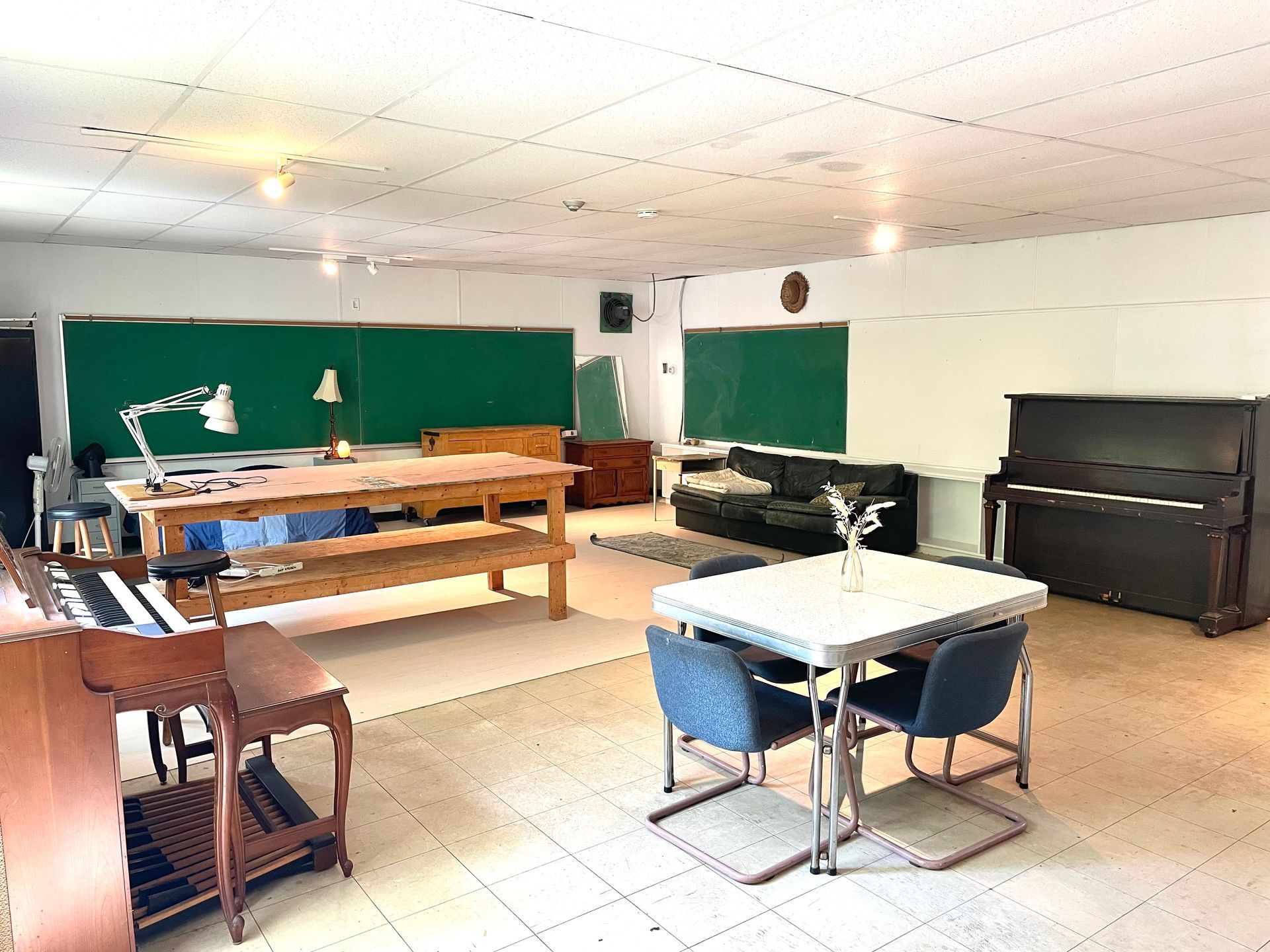 Large, eclectic room with a piano, desks, green chalkboards, a couch, and a table with chairs.