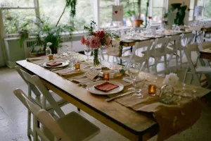 Long wooden table set for a meal with white chairs, flowers, candles, and windows in the background.